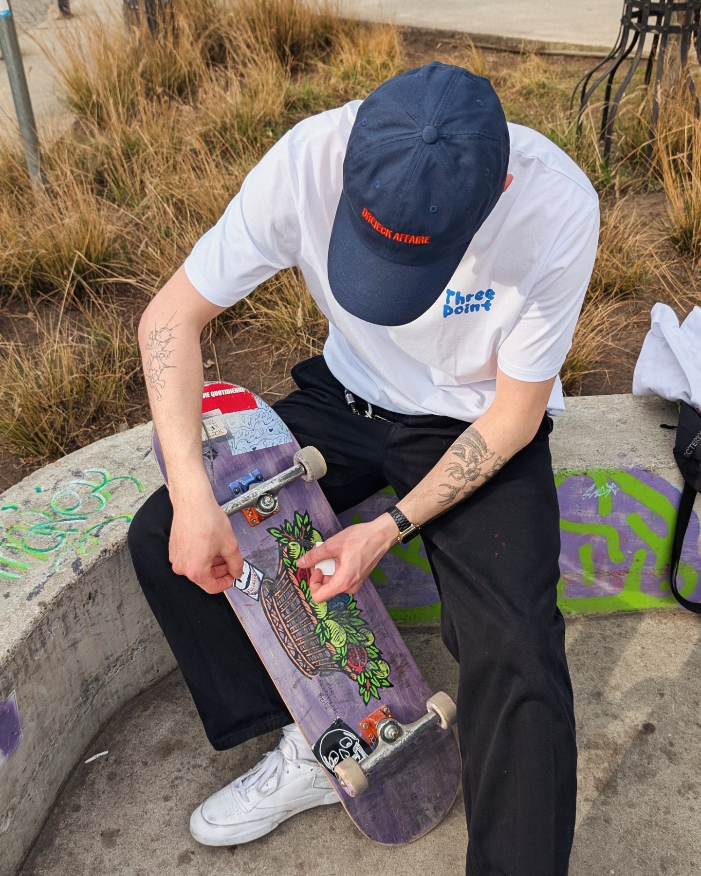 Ein Skateboarder in einem weißen Unisex Bio-Baumwoll-T-Shirt mit dem kleinen "Three Point" Logo auf der Brust sitzt auf einer Betonmauer und wechselt Rollen an seinem Skateboard.