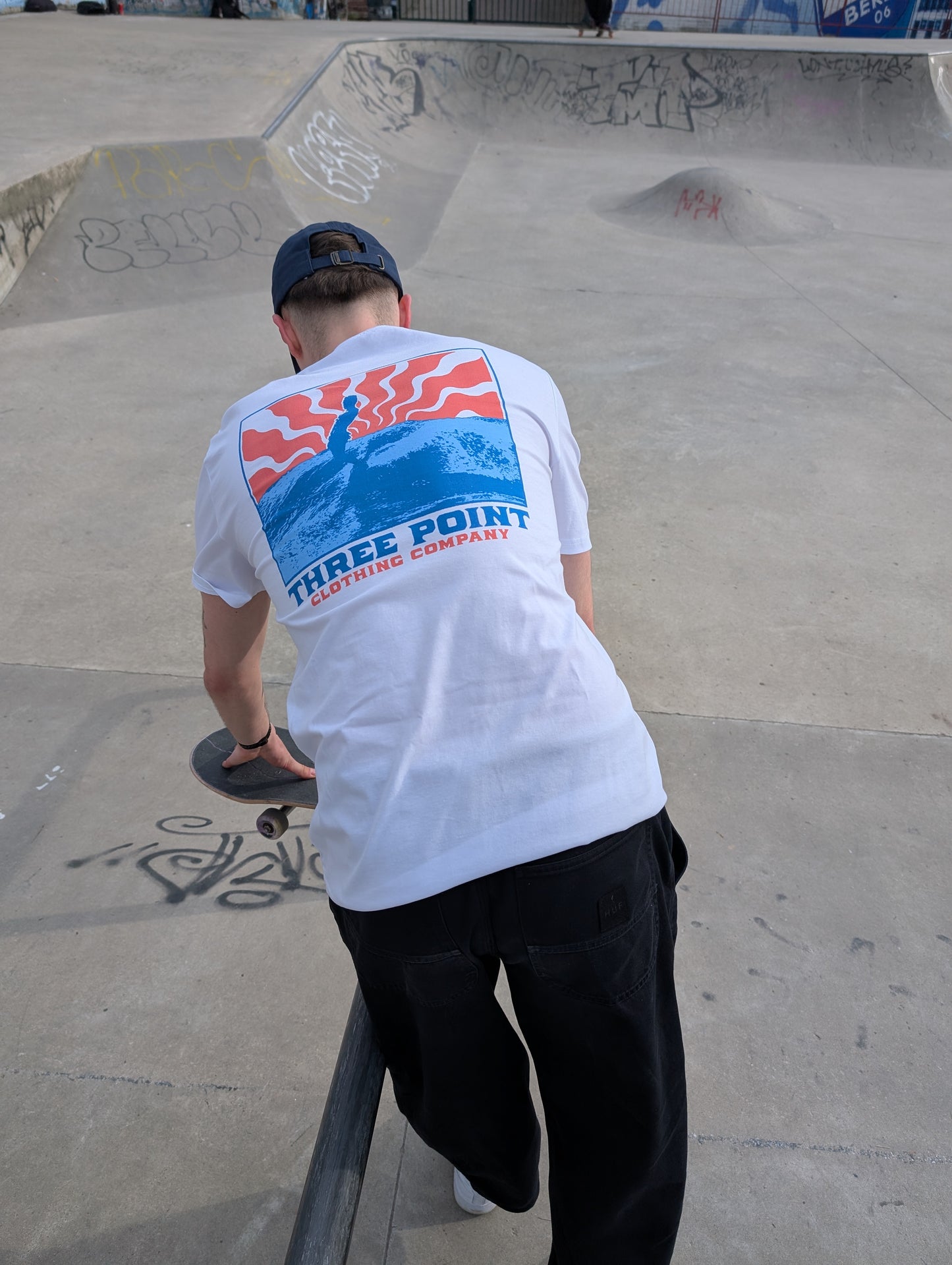 Ein Skateboarder in einem weißen T-Shirt mit einem farbigen "Beton Surfer" Design auf dem Rücken steht mit seinem Skateboard an einer Metallstange in einm Skatepark.Dabei schaut er ob er seine Tricks an der Stange mit Skateboard üben kann.