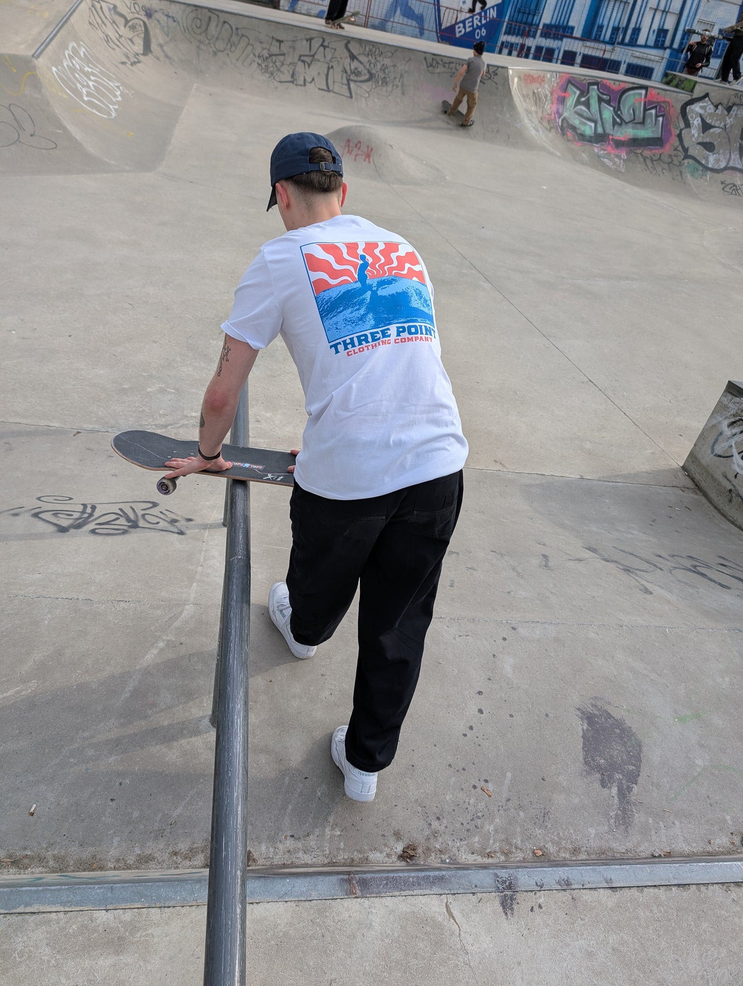 Ein Skateboarder in einem weißen T-Shirt mit einem farbigen "Beton Surfer" Design auf dem Rücken steht mit seinem Skateboard an einer Metallstange in einm Skatepark.